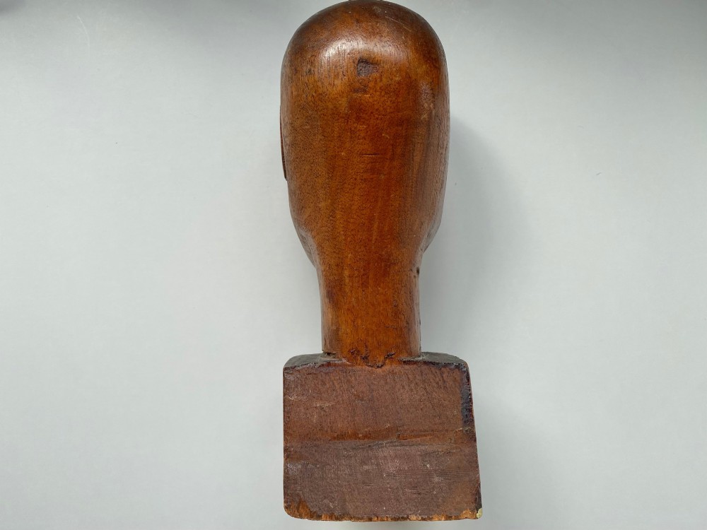 A small hand-carved wooden head sculpture on a square base with stylized eyes and mouth in a warm brown finish. View V