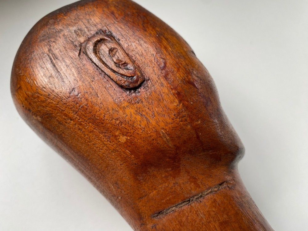 A small hand-carved wooden head sculpture on a square base with stylized eyes and mouth in a warm brown finish. View VI