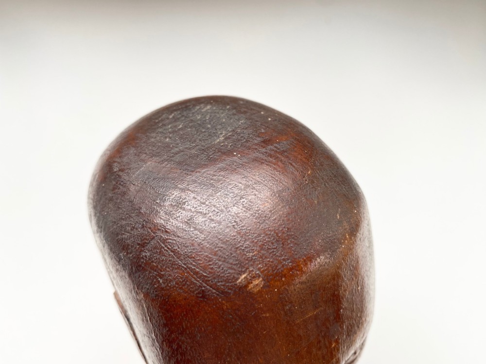 A small hand-carved wooden head sculpture on a square base with stylized eyes and mouth in a warm brown finish. View IX