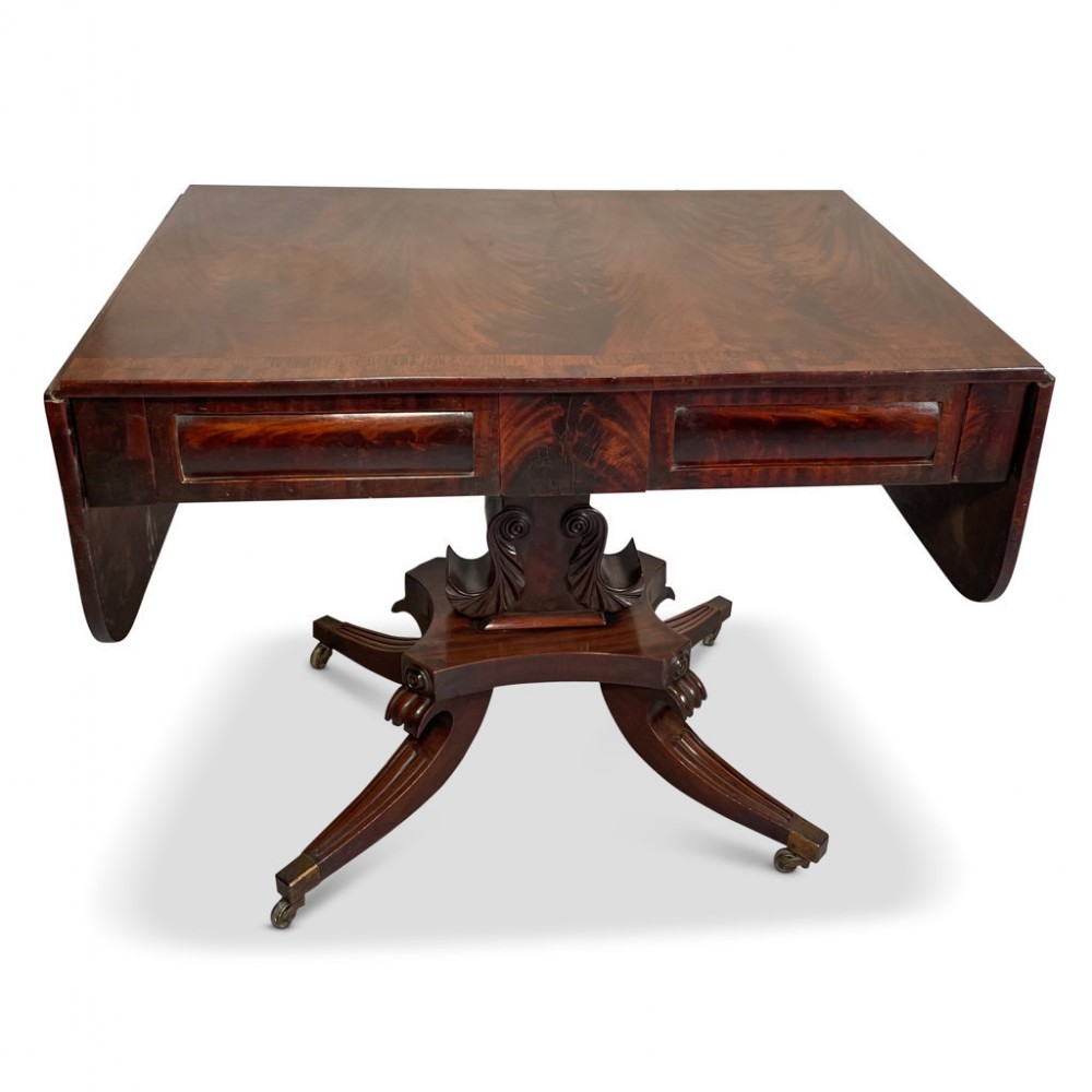 A Regency mahogany sofa table featuring twin drop flaps, two frieze drawers, a carved pedestal column, and four splayed sabre legs with brass castors.