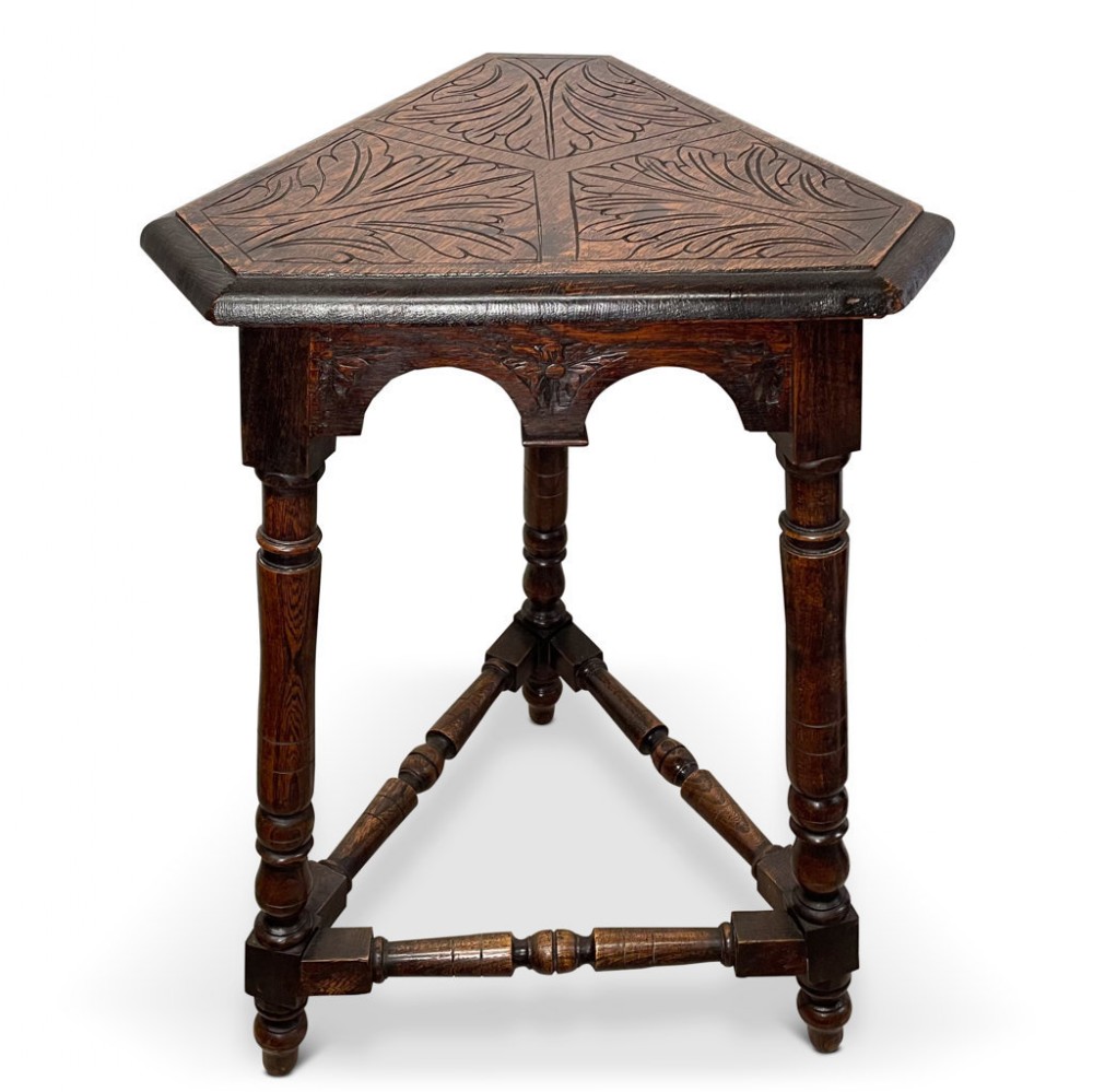 A dark oak three-legged side table with a triangular carved leaf-pattern top, arched aprons, and turned legs connected by stretchers.