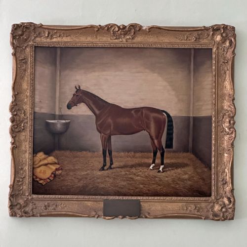 fig 1. An antique Victorian oil painting depicting a bay racehorse standing in profile inside a straw-lined stable stall.