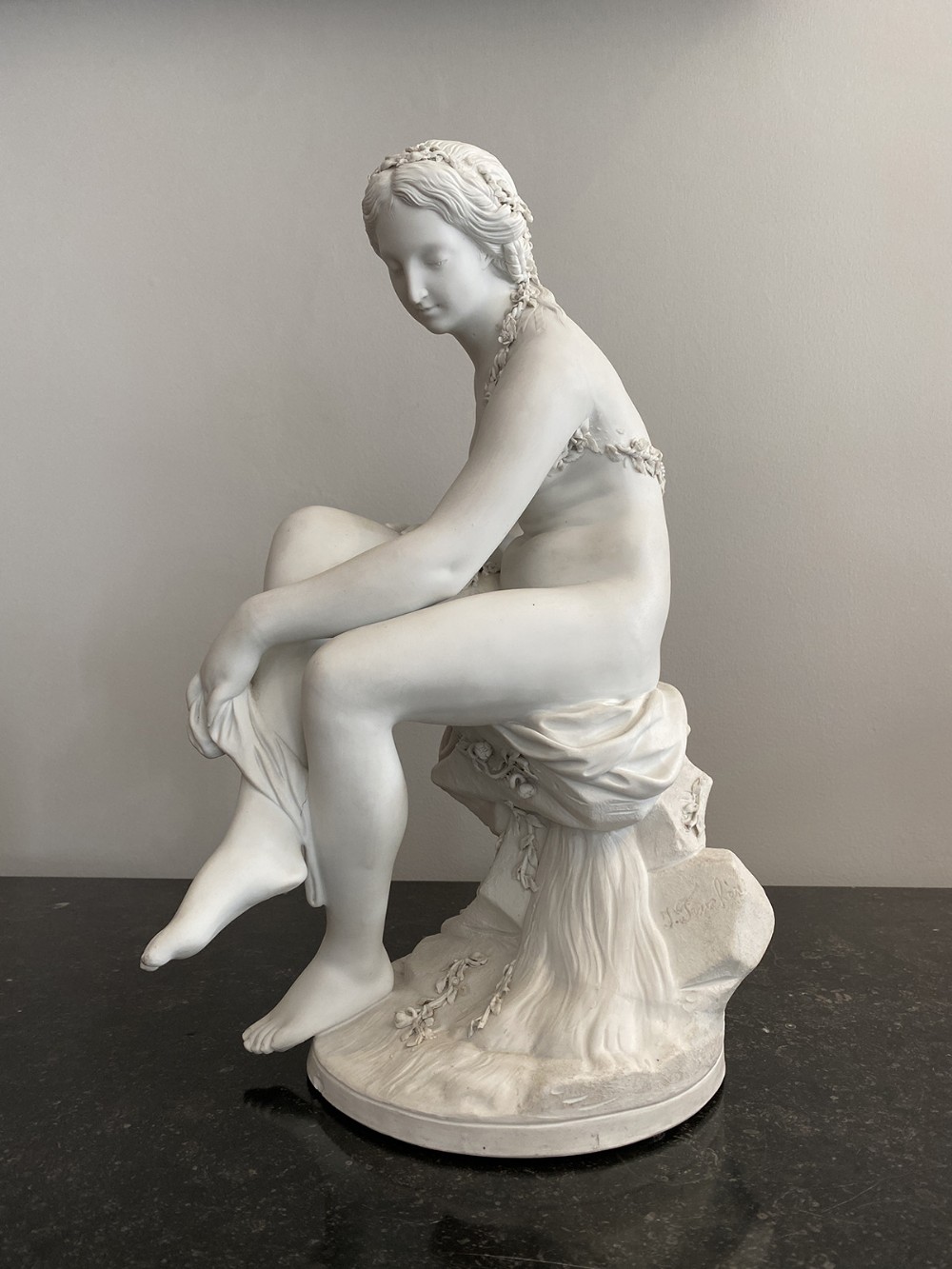 A matte white biscuit porcelain sculpture depicting a seated female nude figure resting upon a naturalistic rocky base.