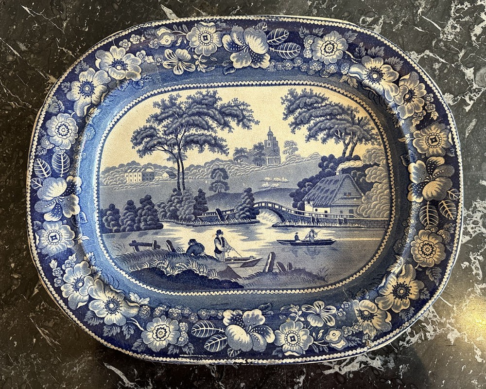 An oval blue and white transferware ceramic platter featuring a detailed pastoral river landscape framed by a wide floral border.