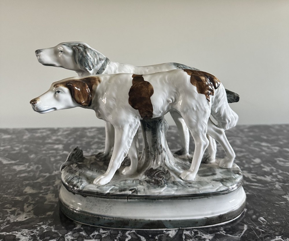 A glazed ceramic figurine featuring a pair of standing dogs with brown and white patches on a naturalistic oval base.