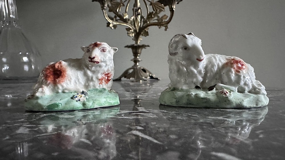 A pair of 18th-century painted porcelain sheep figurines depicting a ram and a ewe reclining on green bases with floral accents.