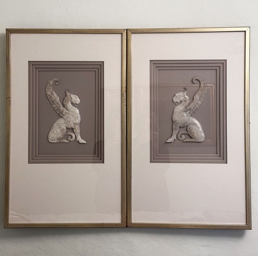 A pair of framed relief sculptures depicting carved stone griffin figures facing each other within layered grey mounts and gold frames.