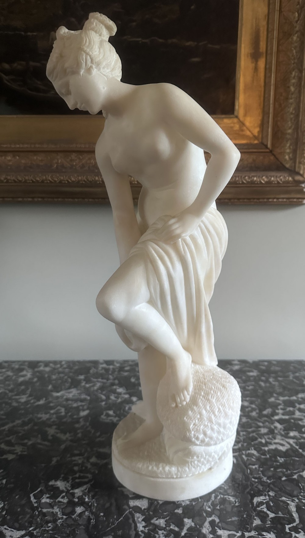 A carved white alabaster statuette depicting a semi-nude classical female bather with draped cloth, standing on a round pedestal.
