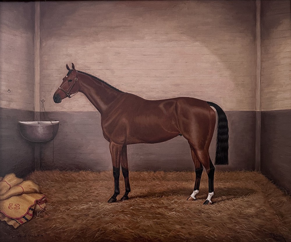 An antique Victorian oil painting depicting a bay racehorse standing in profile inside a straw-lined stable stall.