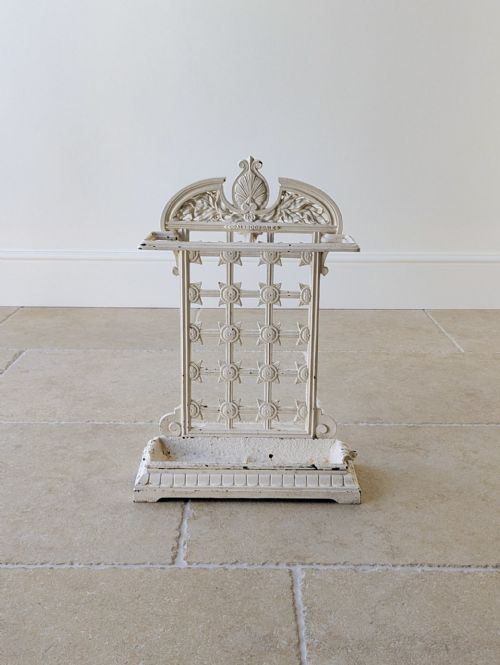 fig 1. An antique off-white painted cast iron stick stand featuring an ornate backplate with shell motifs, a lattice panel, and a rectangular drip tray base.