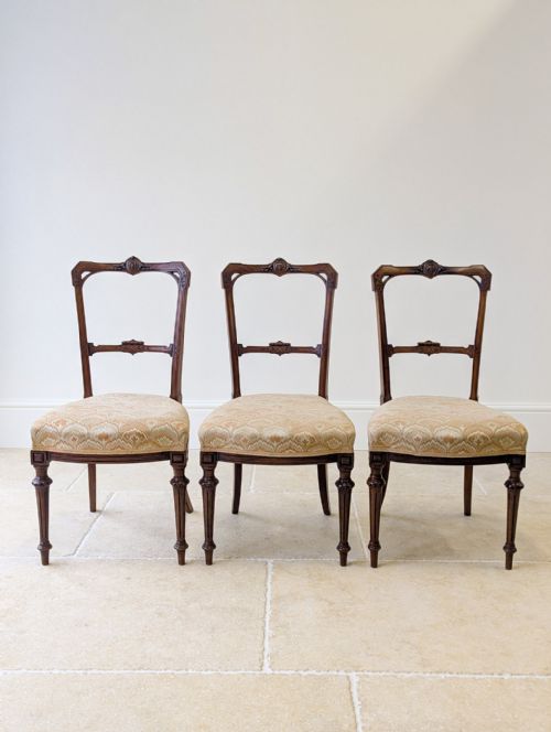 fig 1. A set of three late nineteenth-century Victorian Neoclassical mahogany salon chairs featuring carved crest rails, open backrests, patterned upholstered seats, and fluted front legs.