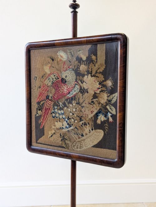 fig 1. A William IV rosewood pole screen featuring a square needlepoint panel displaying a parrot and flowers, set on a turned column with a triform base.