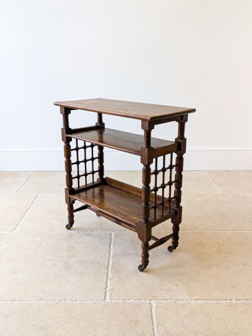 fig 1. A dark oak Arts and Crafts reading table featuring spindle lattice side panels, open tiers, turned legs, and small castors.