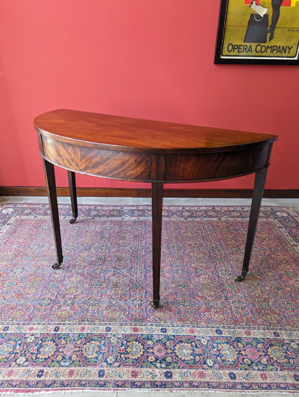 A freestanding antique Victorian mahogany demilune table featuring a curved top, tapered legs with castors, and rich grain patterns.
