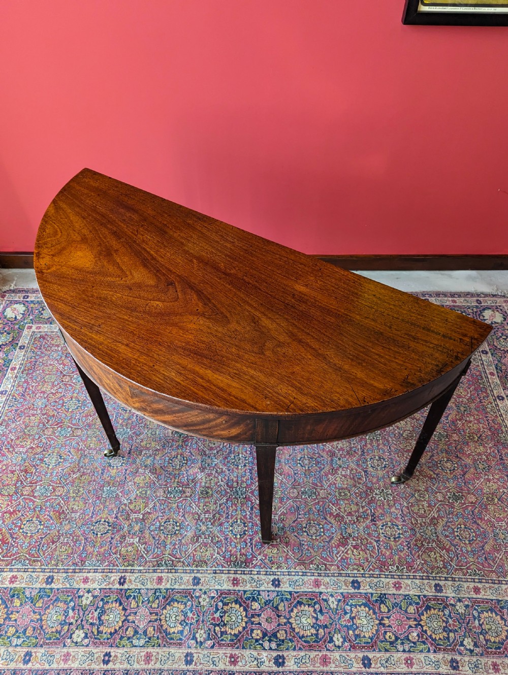 A freestanding antique Victorian mahogany demilune table featuring a curved top, tapered legs with castors, and rich grain patterns. View V