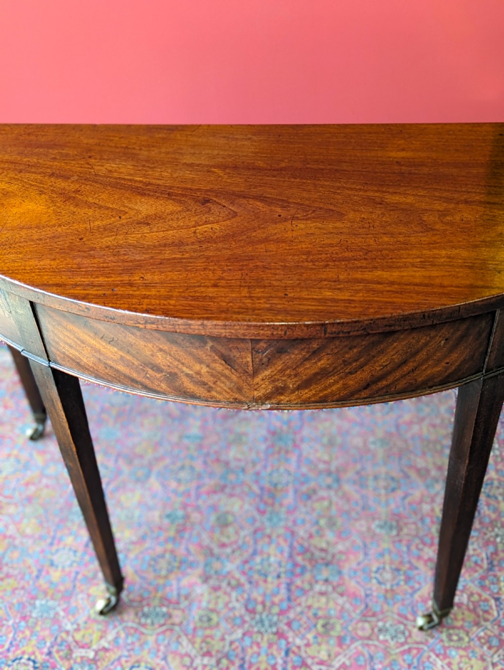 A freestanding antique Victorian mahogany demilune table featuring a curved top, tapered legs with castors, and rich grain patterns. View VI
