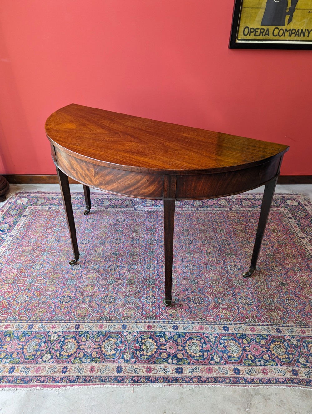 A freestanding antique Victorian mahogany demilune table featuring a curved top, tapered legs with castors, and rich grain patterns. View VIII