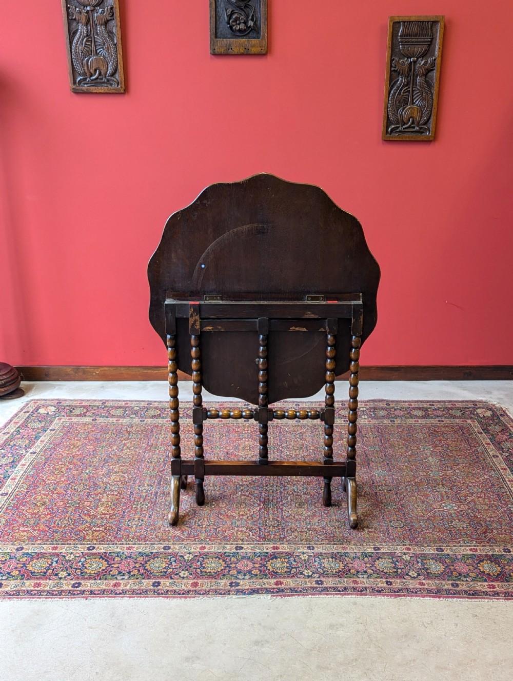 An antique early 20th-century oak folding table featuring a shaped top and turned bobbin legs, shown folded upright. View XI