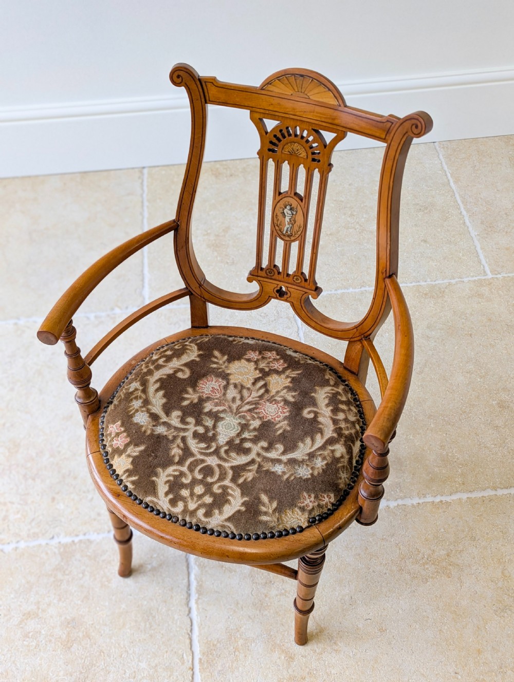 A late Victorian satinwood elbow chair featuring a lyre-shaped backrest with a painted central medallion, turned front legs, and an upholstered seat. View IV