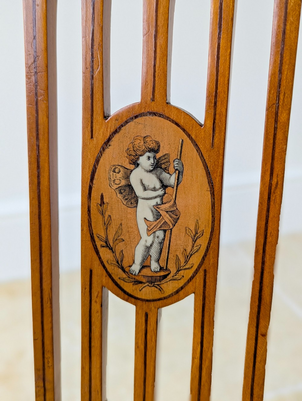 A late Victorian satinwood elbow chair featuring a lyre-shaped backrest with a painted central medallion, turned front legs, and an upholstered seat. View V