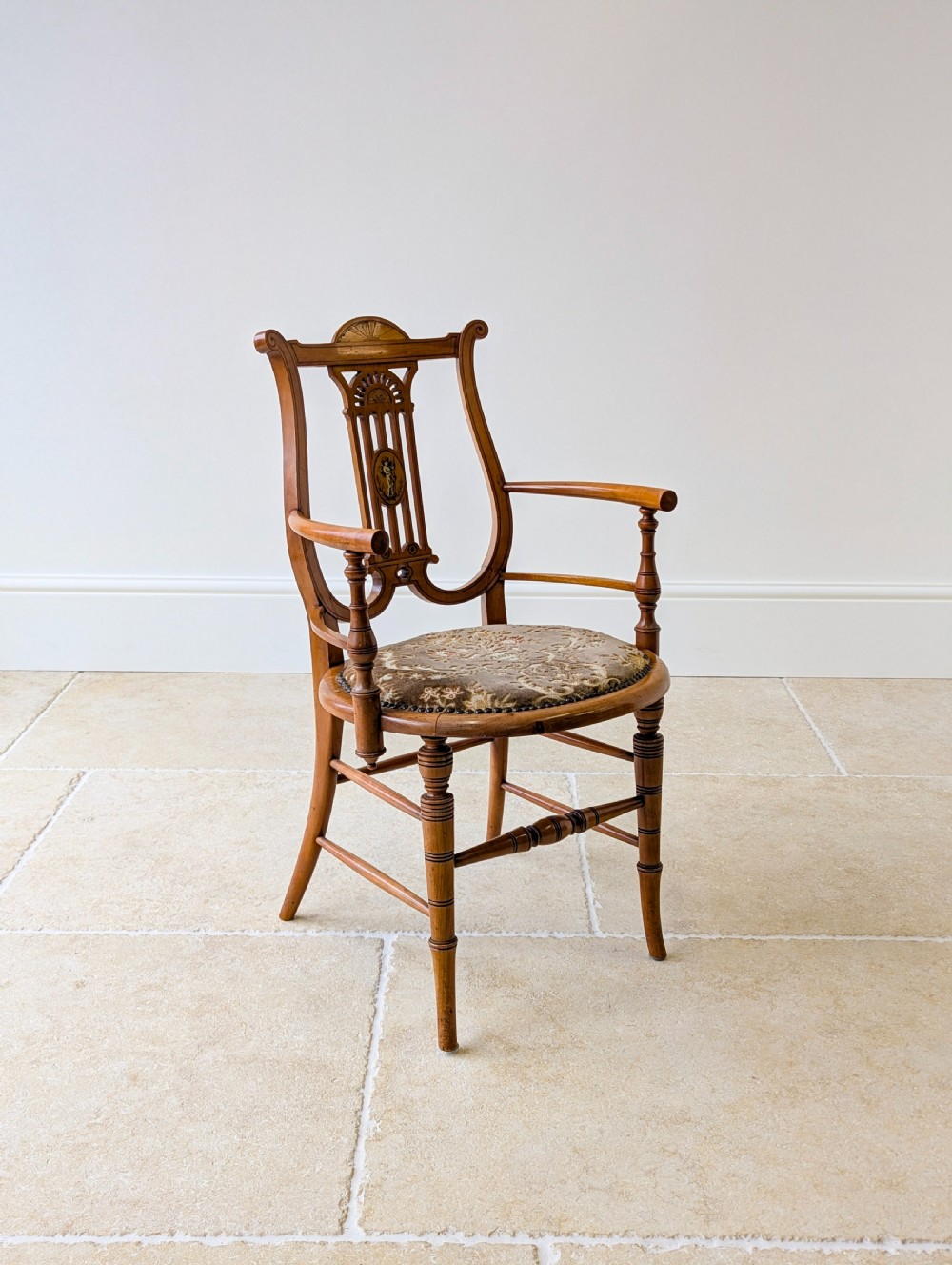A late Victorian satinwood elbow chair featuring a lyre-shaped backrest with a painted central medallion, turned front legs, and an upholstered seat. View VI