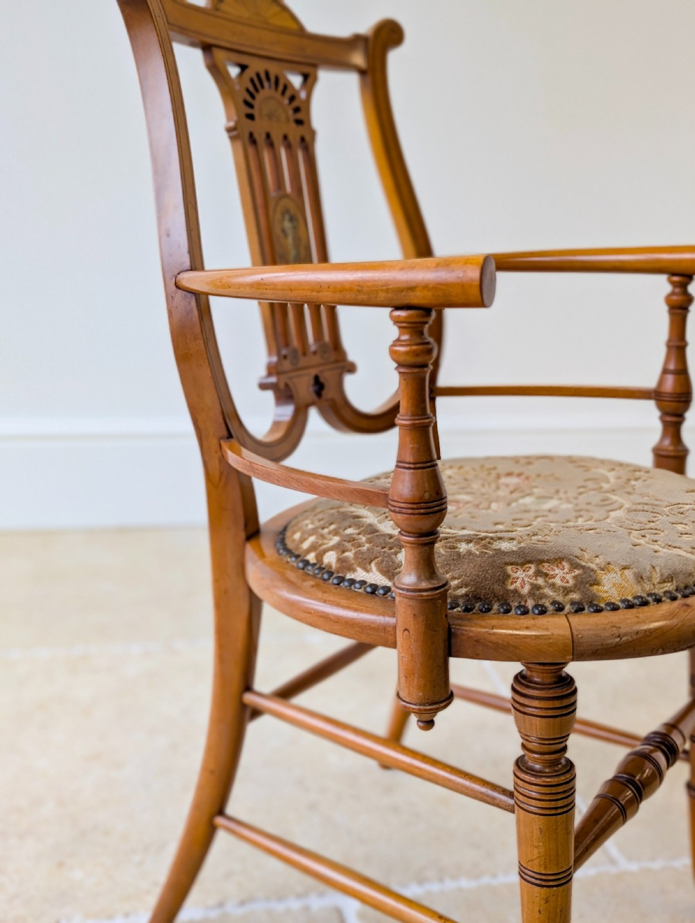 A late Victorian satinwood elbow chair featuring a lyre-shaped backrest with a painted central medallion, turned front legs, and an upholstered seat. View XV