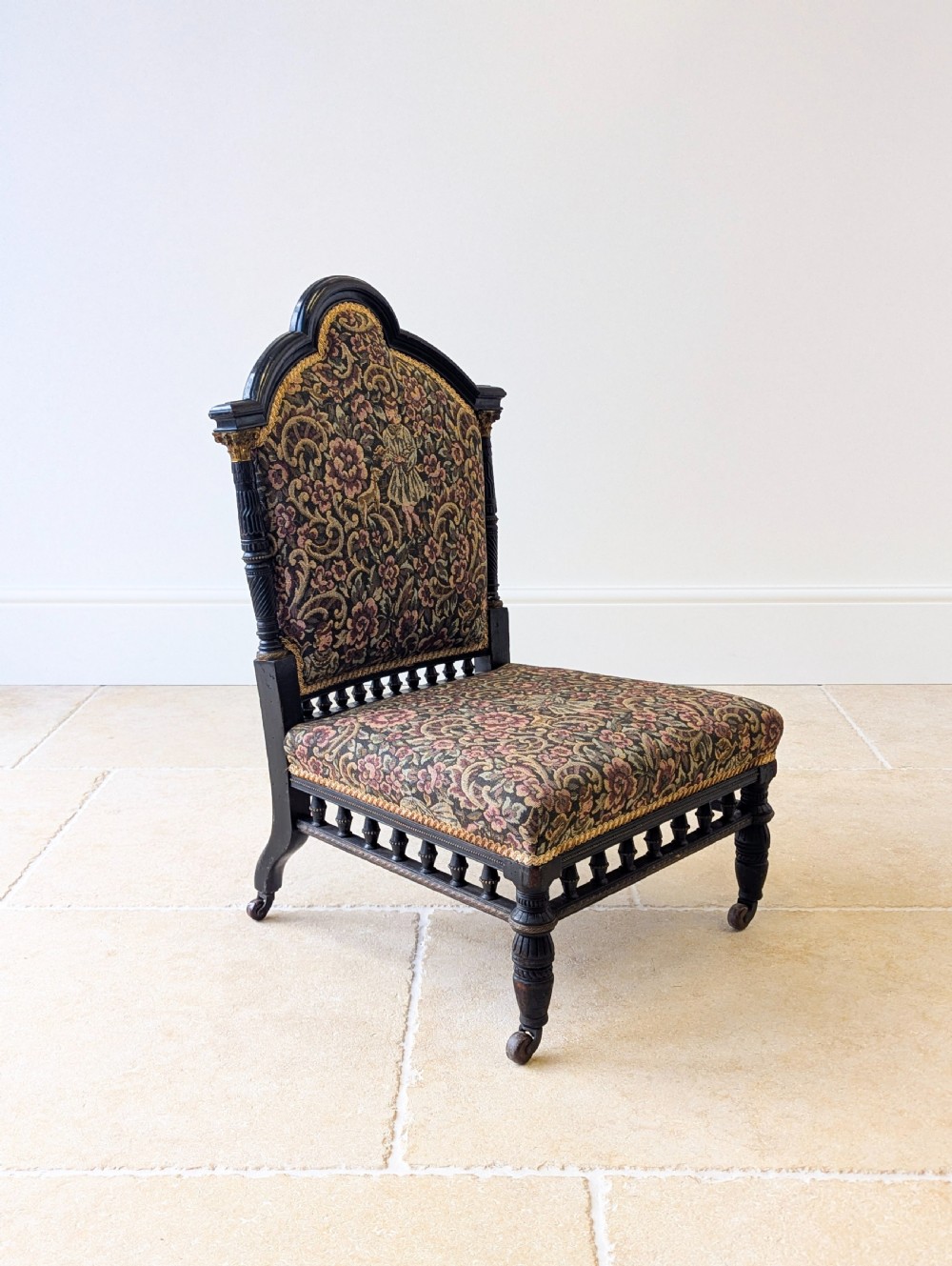 An antique Victorian ebonised low chair featuring a floral tapestry upholstered seat and backrest with turned spindle galleries and castors. View II