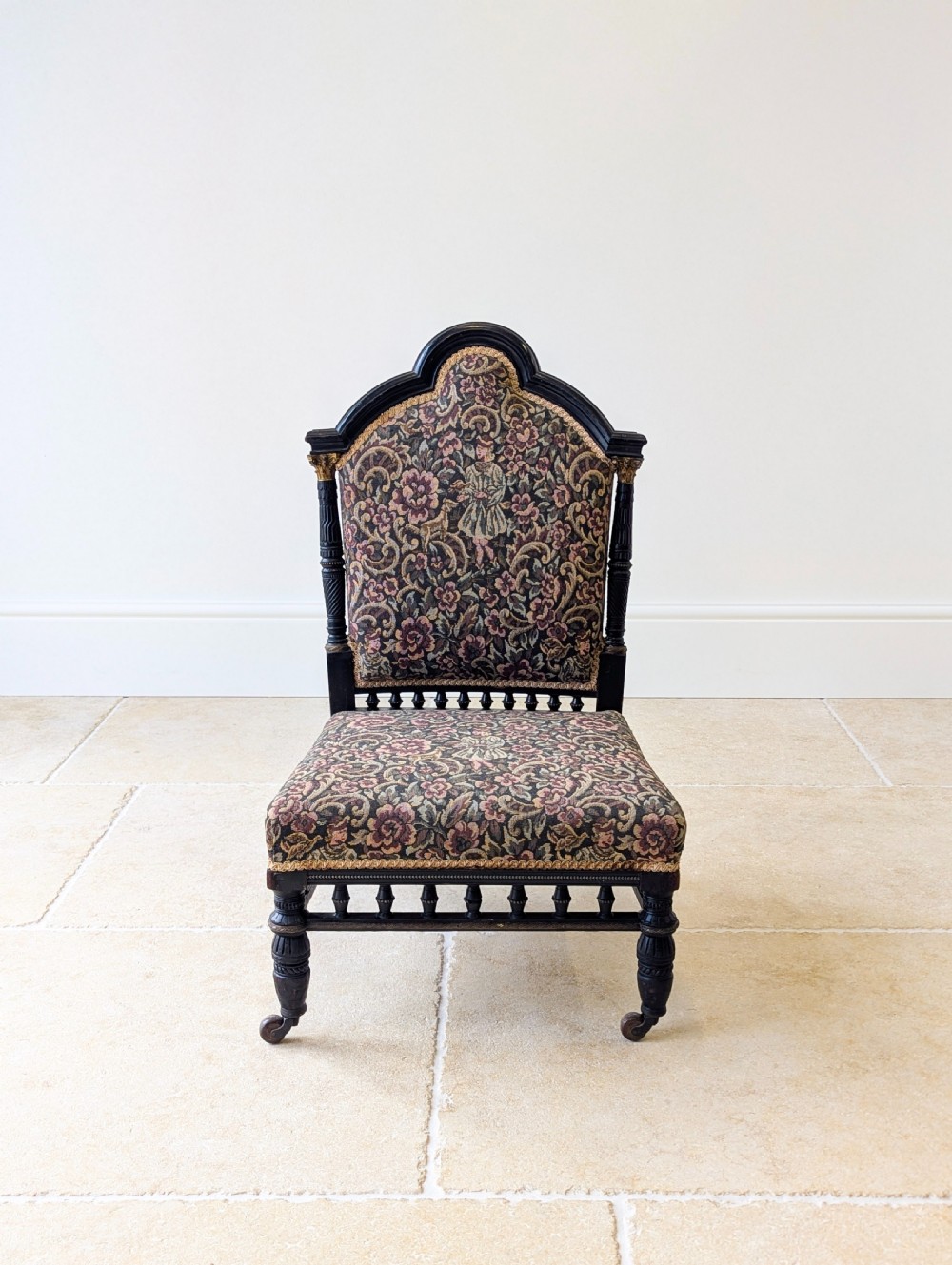 An antique Victorian ebonised low chair featuring a floral tapestry upholstered seat and backrest with turned spindle galleries and castors. View III