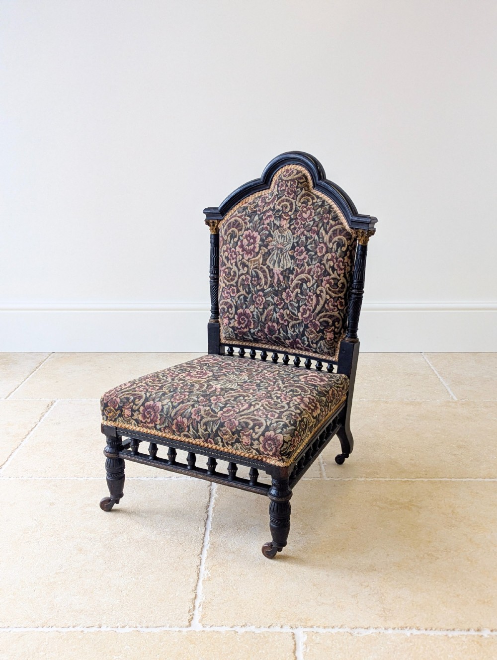 An antique Victorian ebonised low chair featuring a floral tapestry upholstered seat and backrest with turned spindle galleries and castors. View V