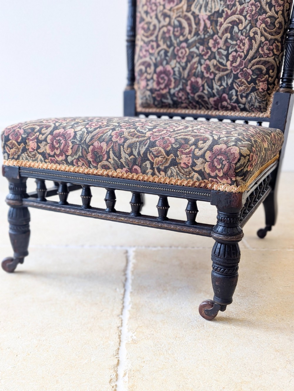 An antique Victorian ebonised low chair featuring a floral tapestry upholstered seat and backrest with turned spindle galleries and castors. View VI