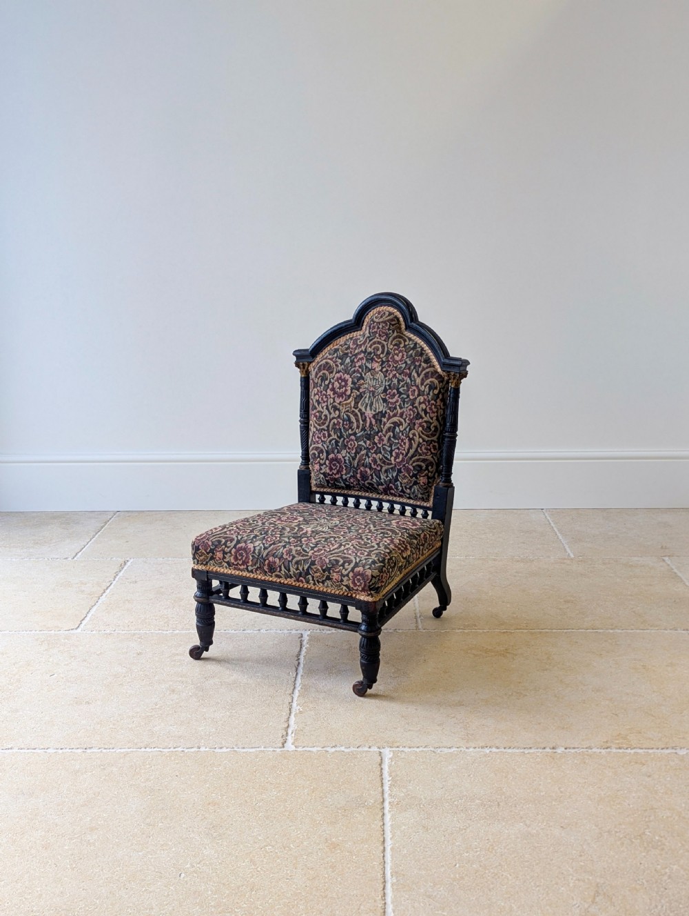 An antique Victorian ebonised low chair featuring a floral tapestry upholstered seat and backrest with turned spindle galleries and castors. View VII