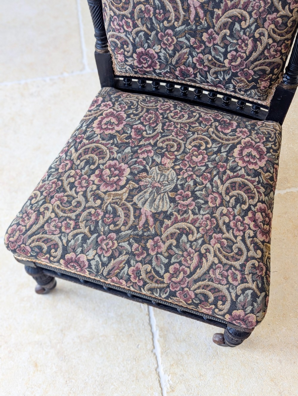 An antique Victorian ebonised low chair featuring a floral tapestry upholstered seat and backrest with turned spindle galleries and castors. View VIII