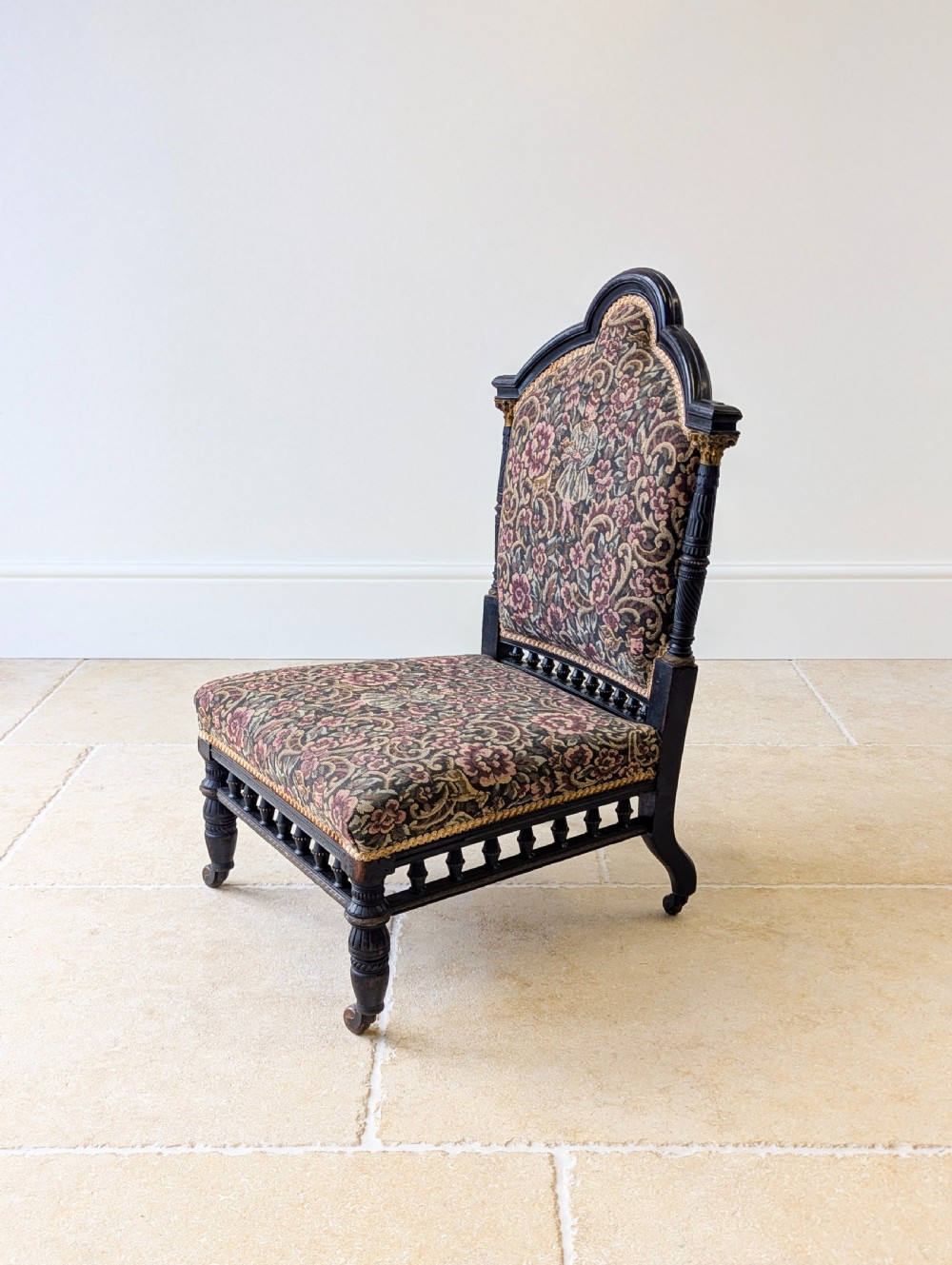 An antique Victorian ebonised low chair featuring a floral tapestry upholstered seat and backrest with turned spindle galleries and castors. View IX