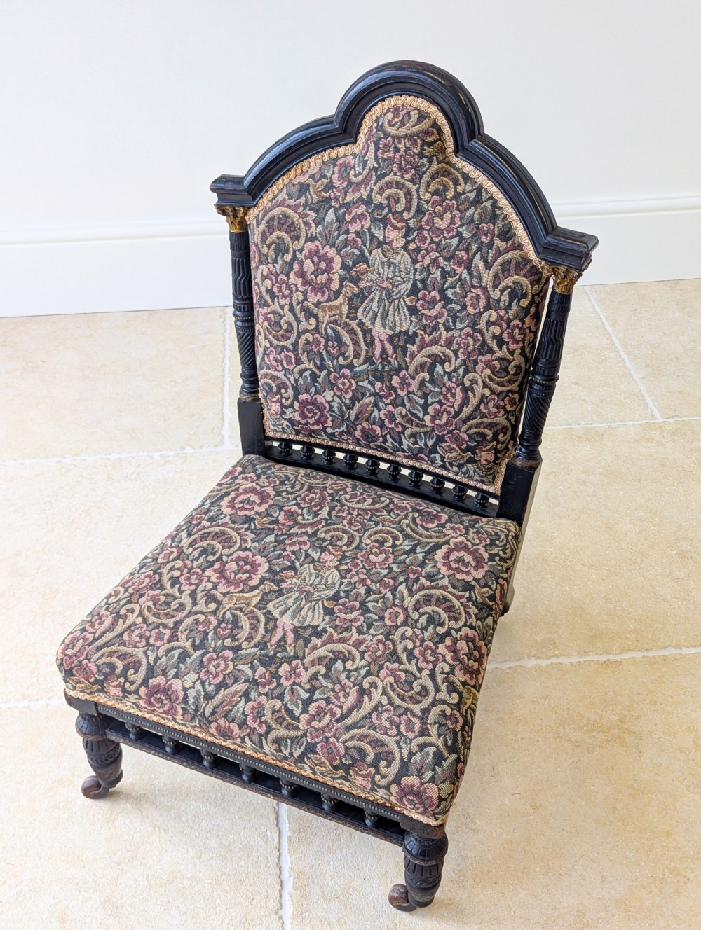 An antique Victorian ebonised low chair featuring a floral tapestry upholstered seat and backrest with turned spindle galleries and castors. View X