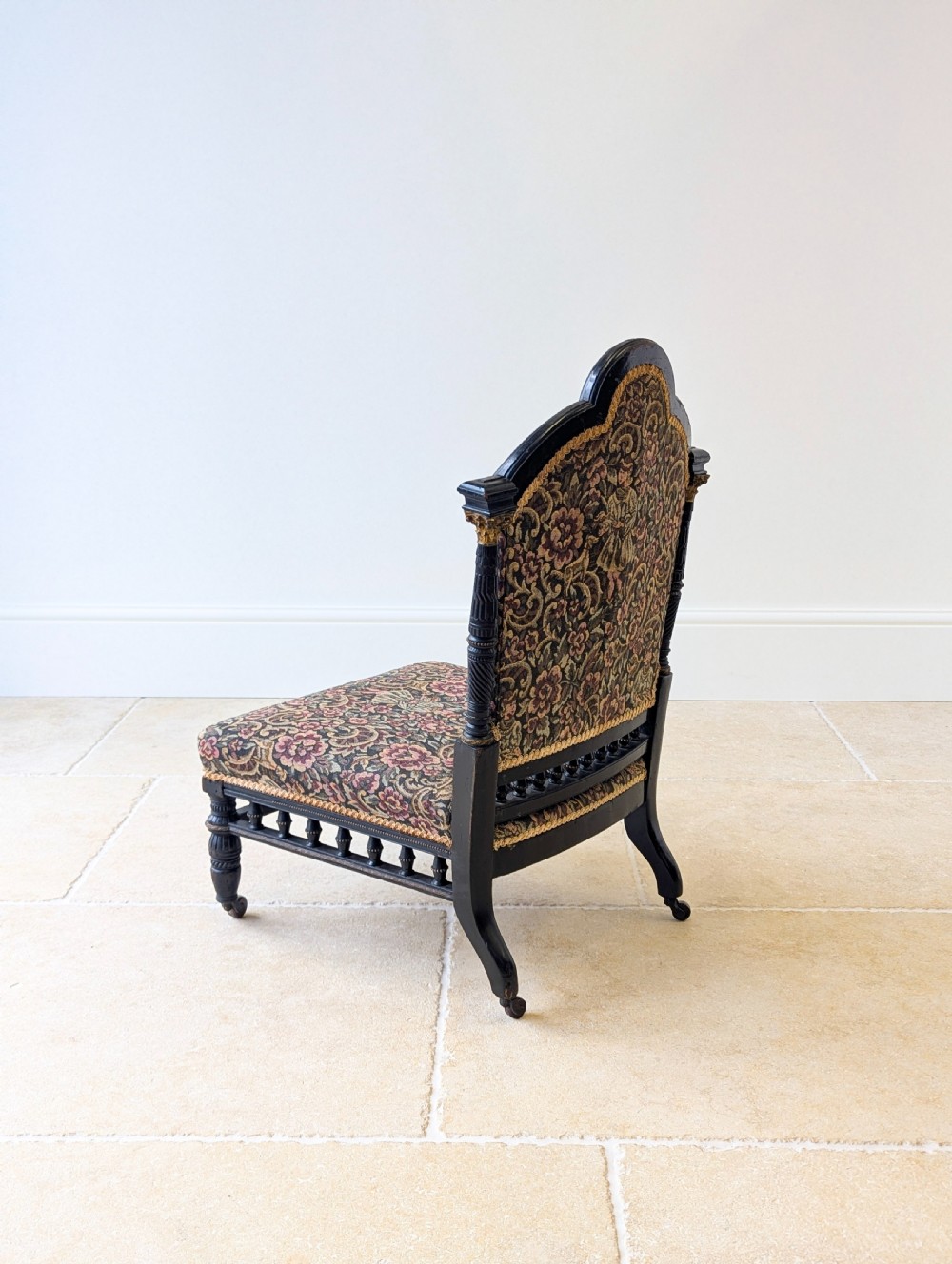 An antique Victorian ebonised low chair featuring a floral tapestry upholstered seat and backrest with turned spindle galleries and castors. View XI