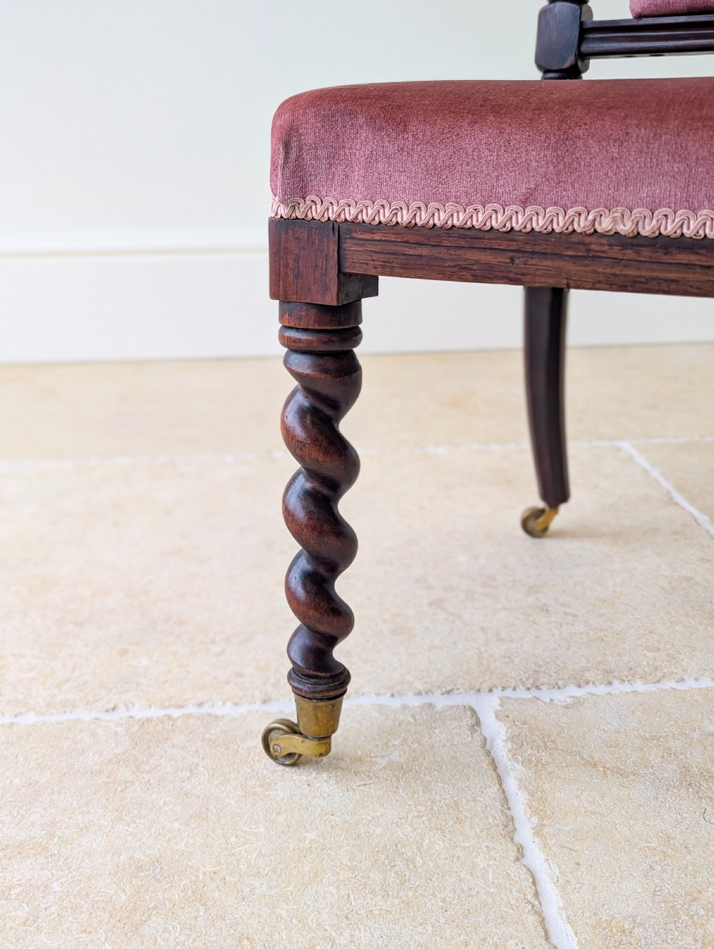 A Victorian rosewood side chair featuring barley twist legs and uprights, a carved cresting rail, dusty pink velvet upholstery, and brass castors. View VI