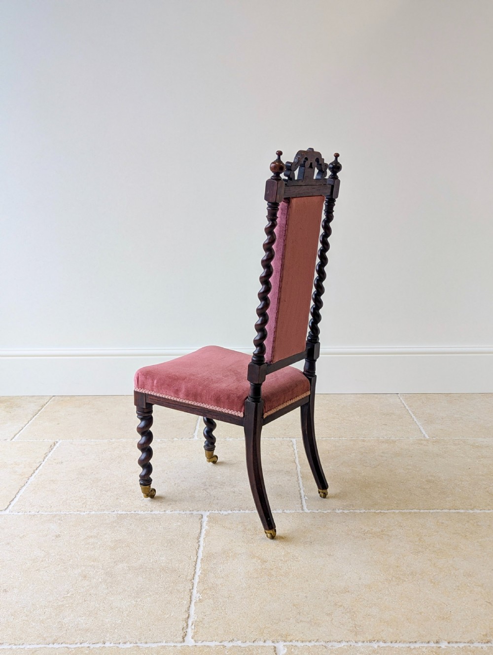 A Victorian rosewood side chair featuring barley twist legs and uprights, a carved cresting rail, dusty pink velvet upholstery, and brass castors. View XV