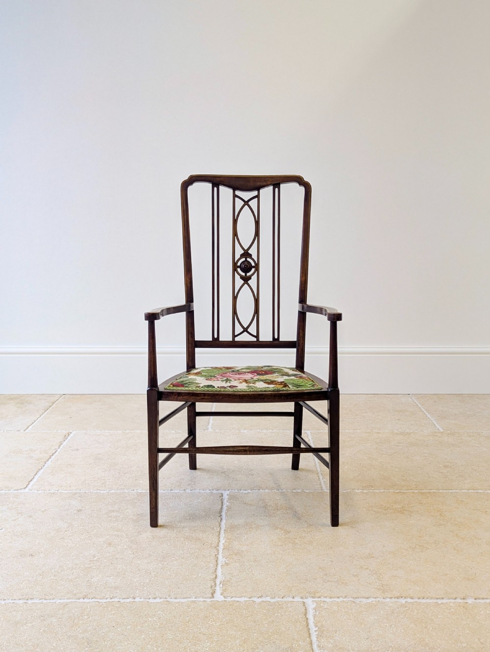 An Edwardian dark mahogany elbow chair featuring a tall pierced splat back, sloped armrests, straight legs with stretchers, and a floral tapestry seat.