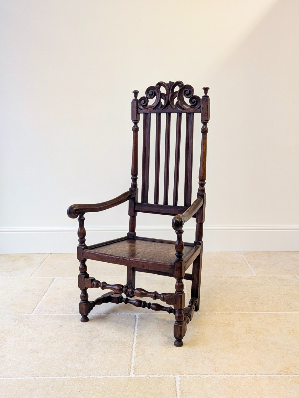 A dark oak William and Mary armchair featuring a carved scrollwork cresting rail, vertical back splats, open arms, and turned legs with stretchers.