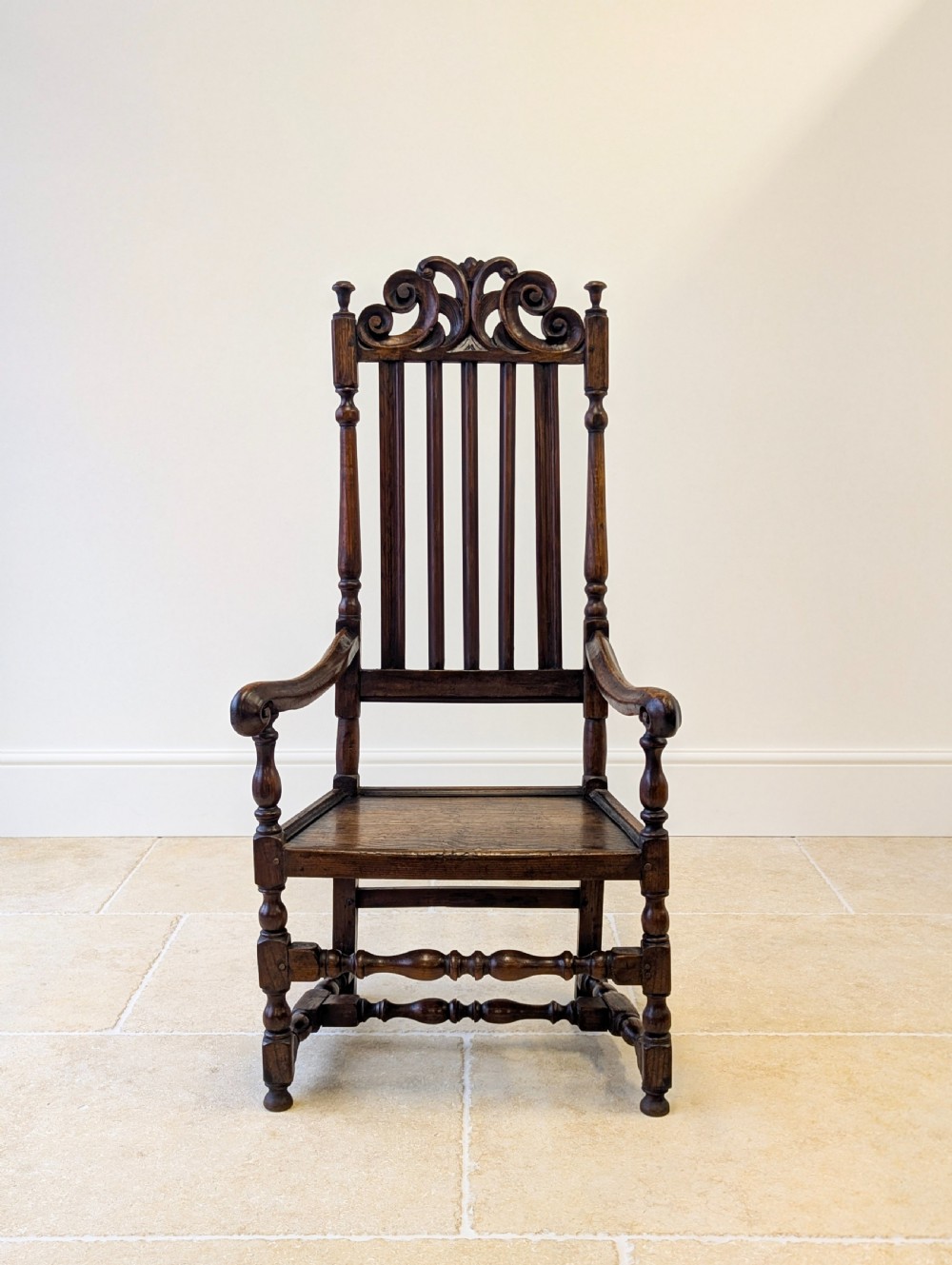 A dark oak William and Mary armchair featuring a carved scrollwork cresting rail, vertical back splats, open arms, and turned legs with stretchers. View II
