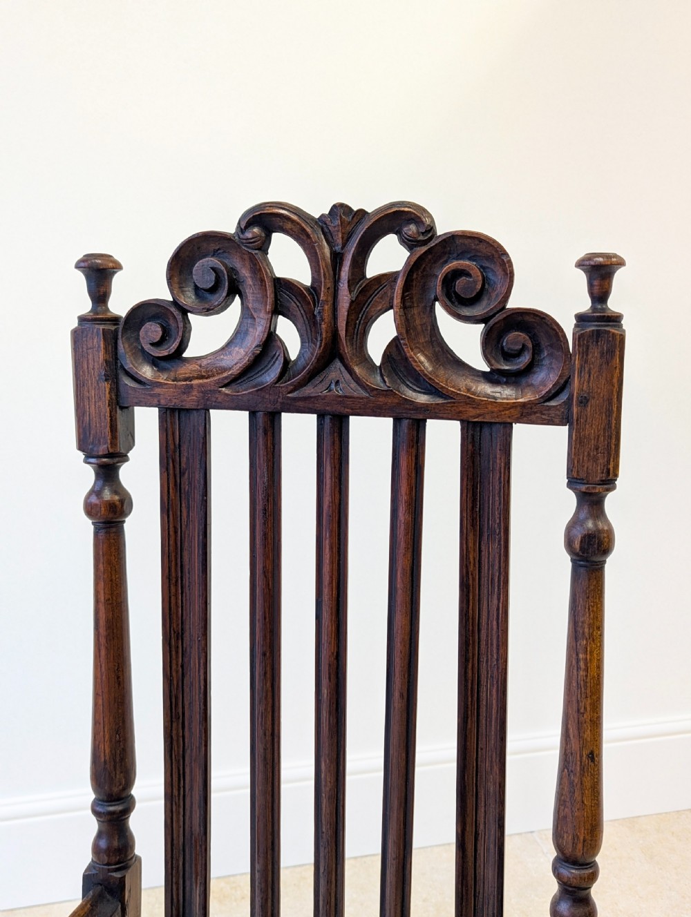 A dark oak William and Mary armchair featuring a carved scrollwork cresting rail, vertical back splats, open arms, and turned legs with stretchers. View III