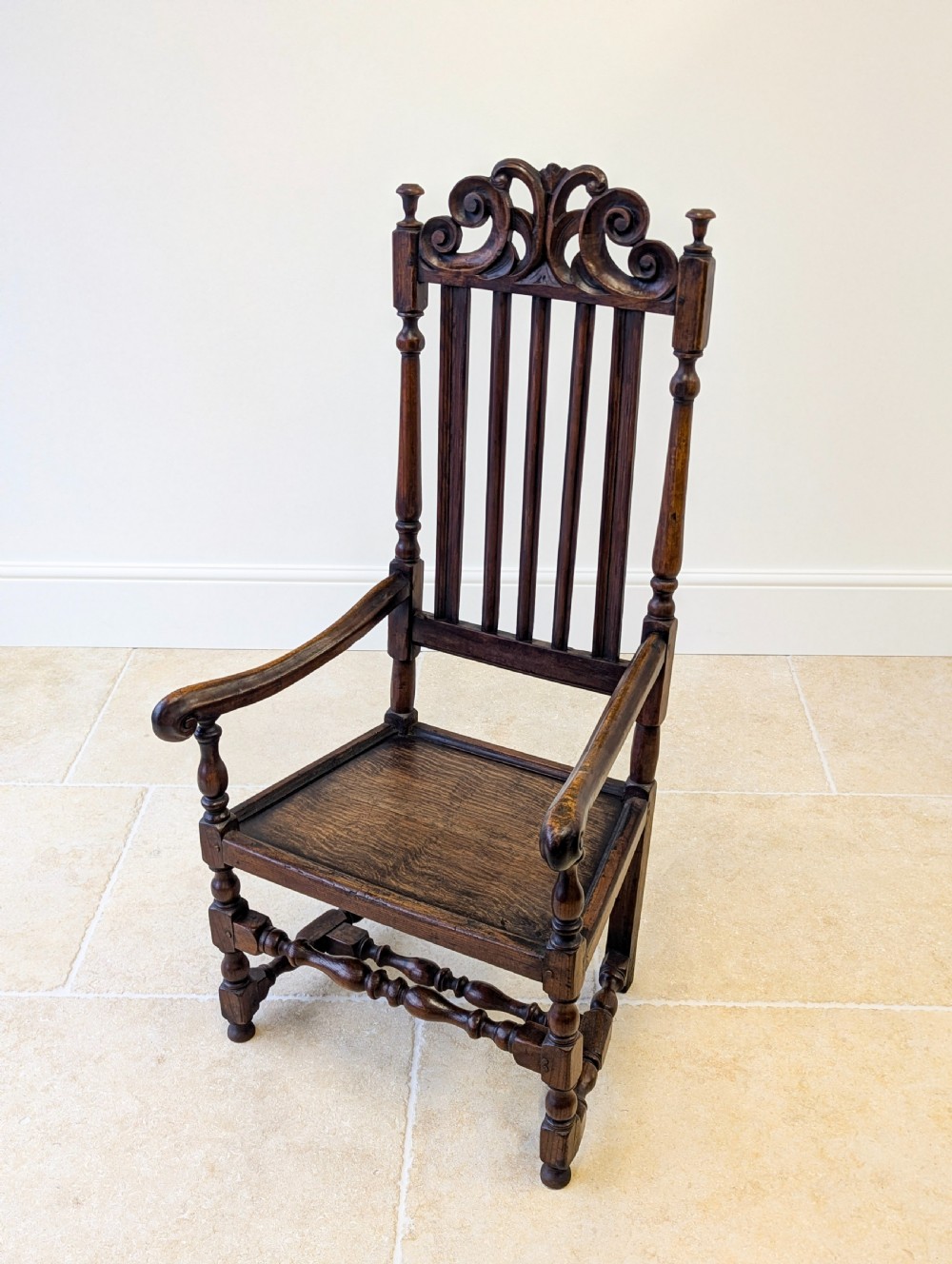 A dark oak William and Mary armchair featuring a carved scrollwork cresting rail, vertical back splats, open arms, and turned legs with stretchers. View IV