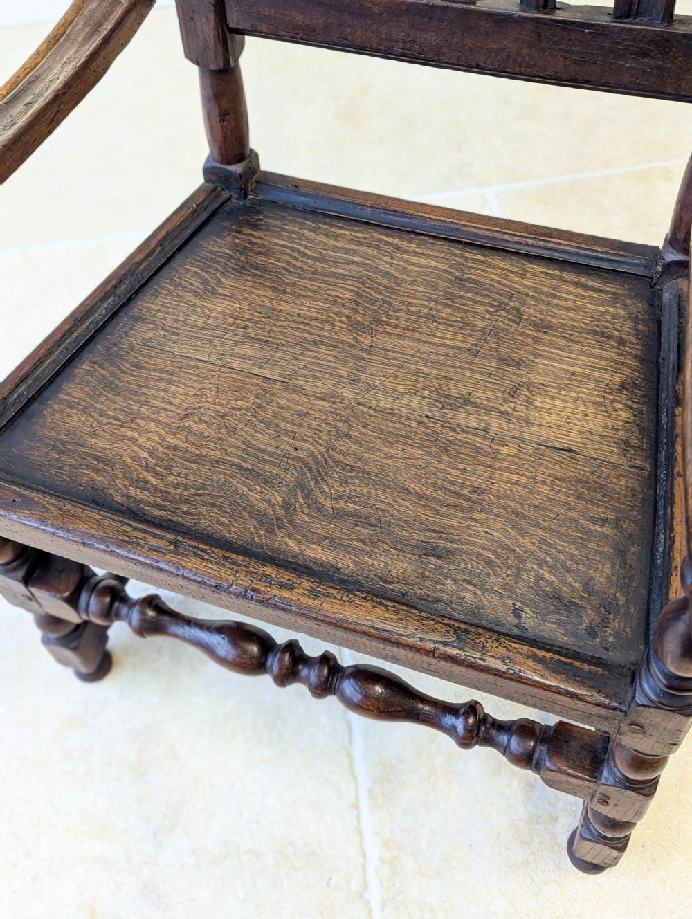 A dark oak William and Mary armchair featuring a carved scrollwork cresting rail, vertical back splats, open arms, and turned legs with stretchers. View V