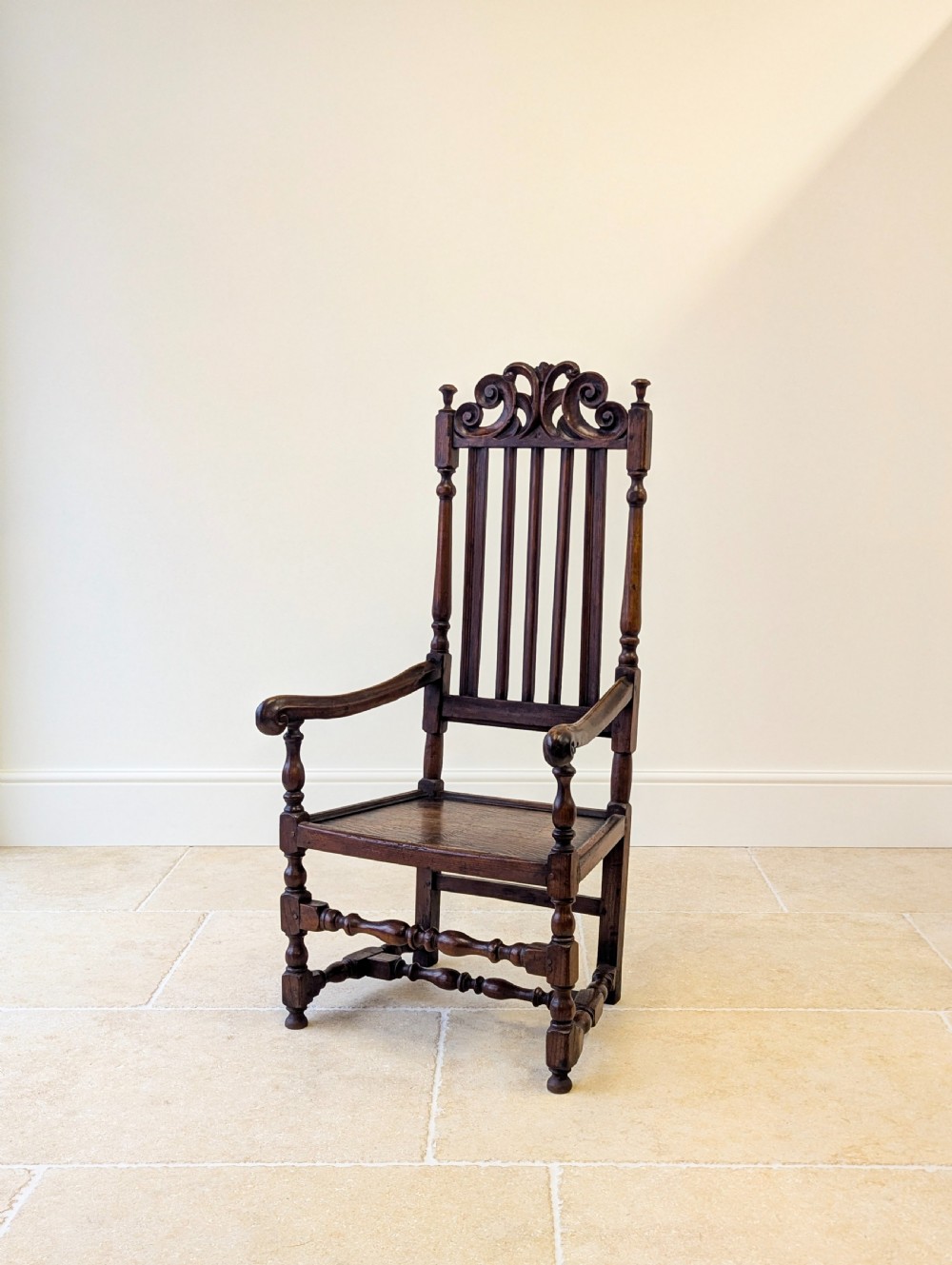 A dark oak William and Mary armchair featuring a carved scrollwork cresting rail, vertical back splats, open arms, and turned legs with stretchers. View VI