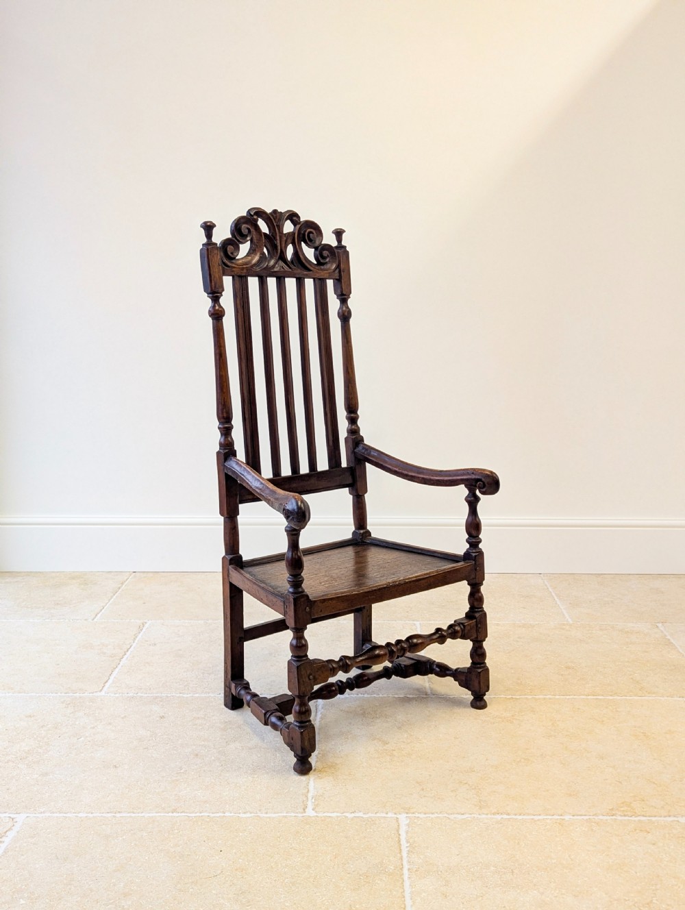 A dark oak William and Mary armchair featuring a carved scrollwork cresting rail, vertical back splats, open arms, and turned legs with stretchers. View X