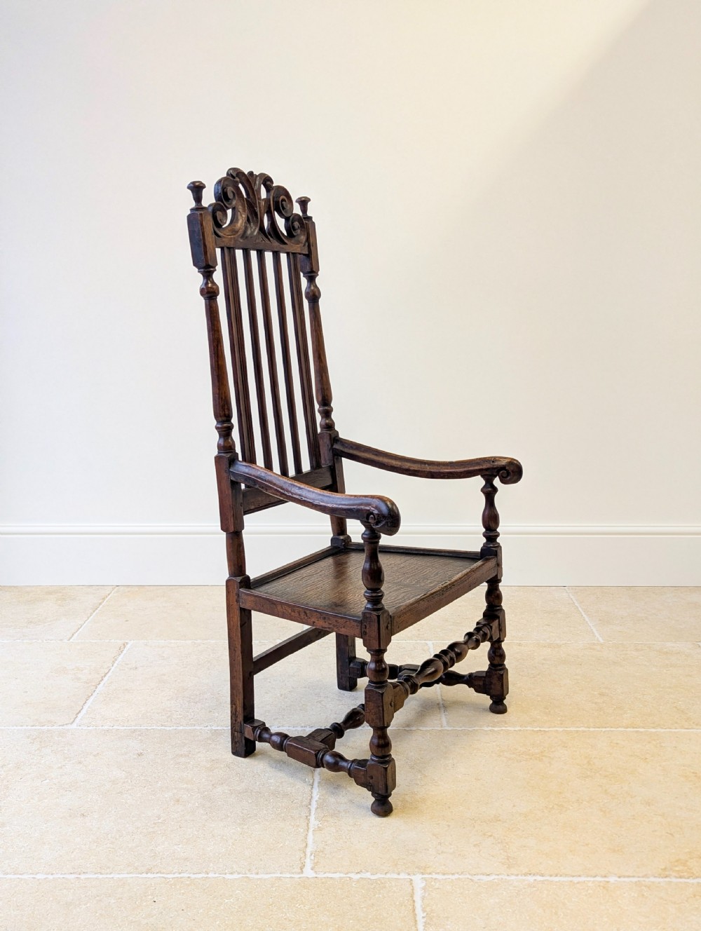 A dark oak William and Mary armchair featuring a carved scrollwork cresting rail, vertical back splats, open arms, and turned legs with stretchers. View XII