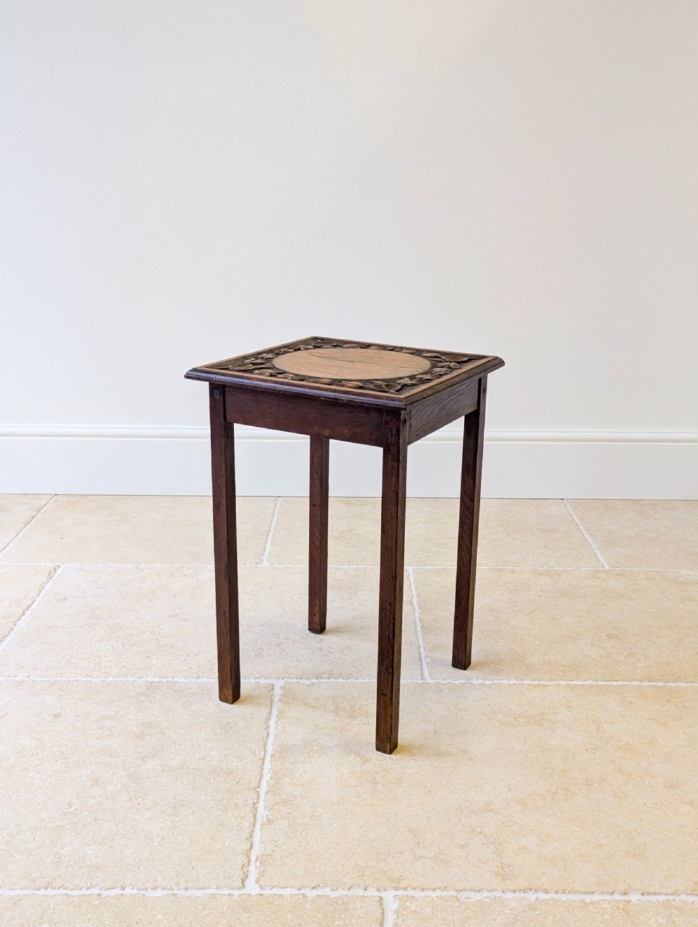A square dark oak occasional table featuring a relief-carved floral border around a central circular panel, supported by four straight square legs.