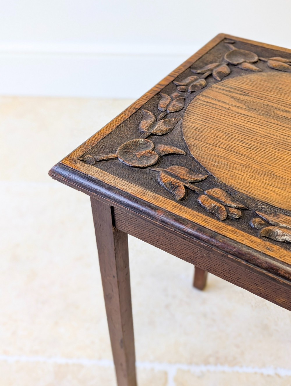 A square dark oak occasional table featuring a relief-carved floral border around a central circular panel, supported by four straight square legs. View IV