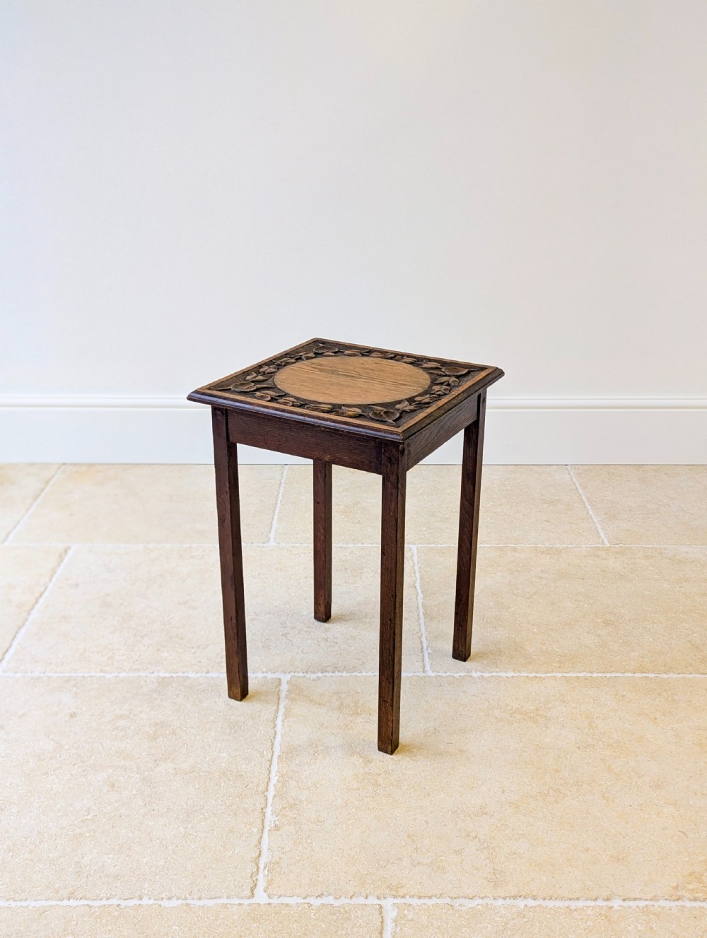 A square dark oak occasional table featuring a relief-carved floral border around a central circular panel, supported by four straight square legs. View V