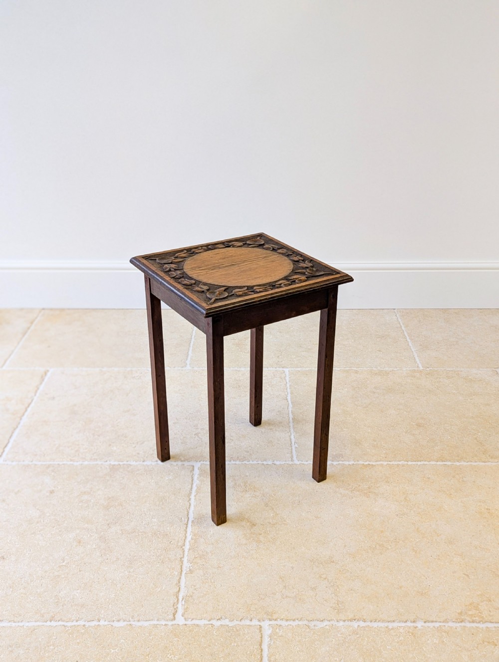 A square dark oak occasional table featuring a relief-carved floral border around a central circular panel, supported by four straight square legs. View IX